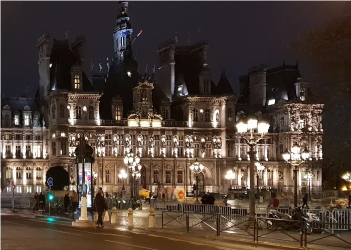 Façade d'un grand bâtiment ancien illuminée la nuit, bordée de lampadaires allumés