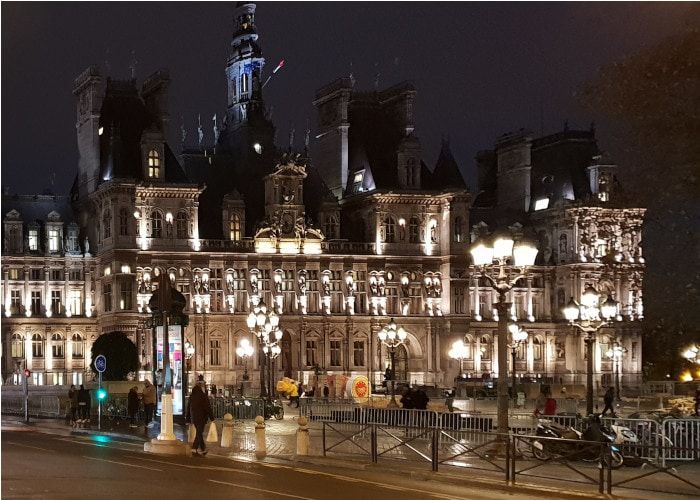 Façade d'un grand bâtiment ancien illuminée la nuit, bordée de lampadaires allumés