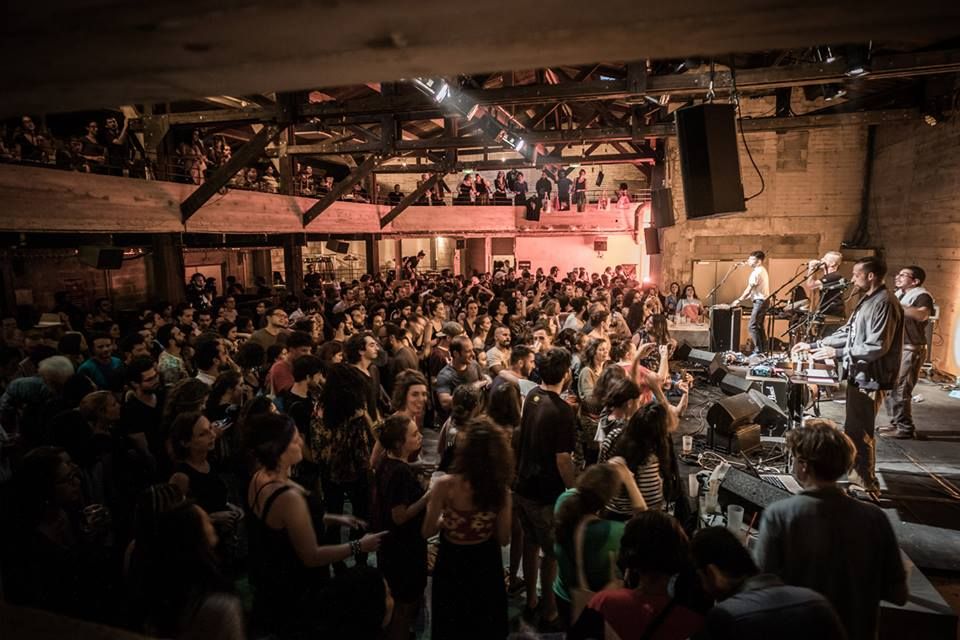 Foule de spectateurs lors d'un concert dans une salle industrielle, avec un groupe sur scène à droite